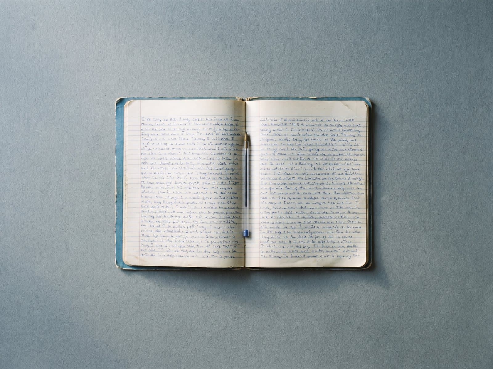 An open school exercise book filled with handwriting in blue ink, a biro in its fold.