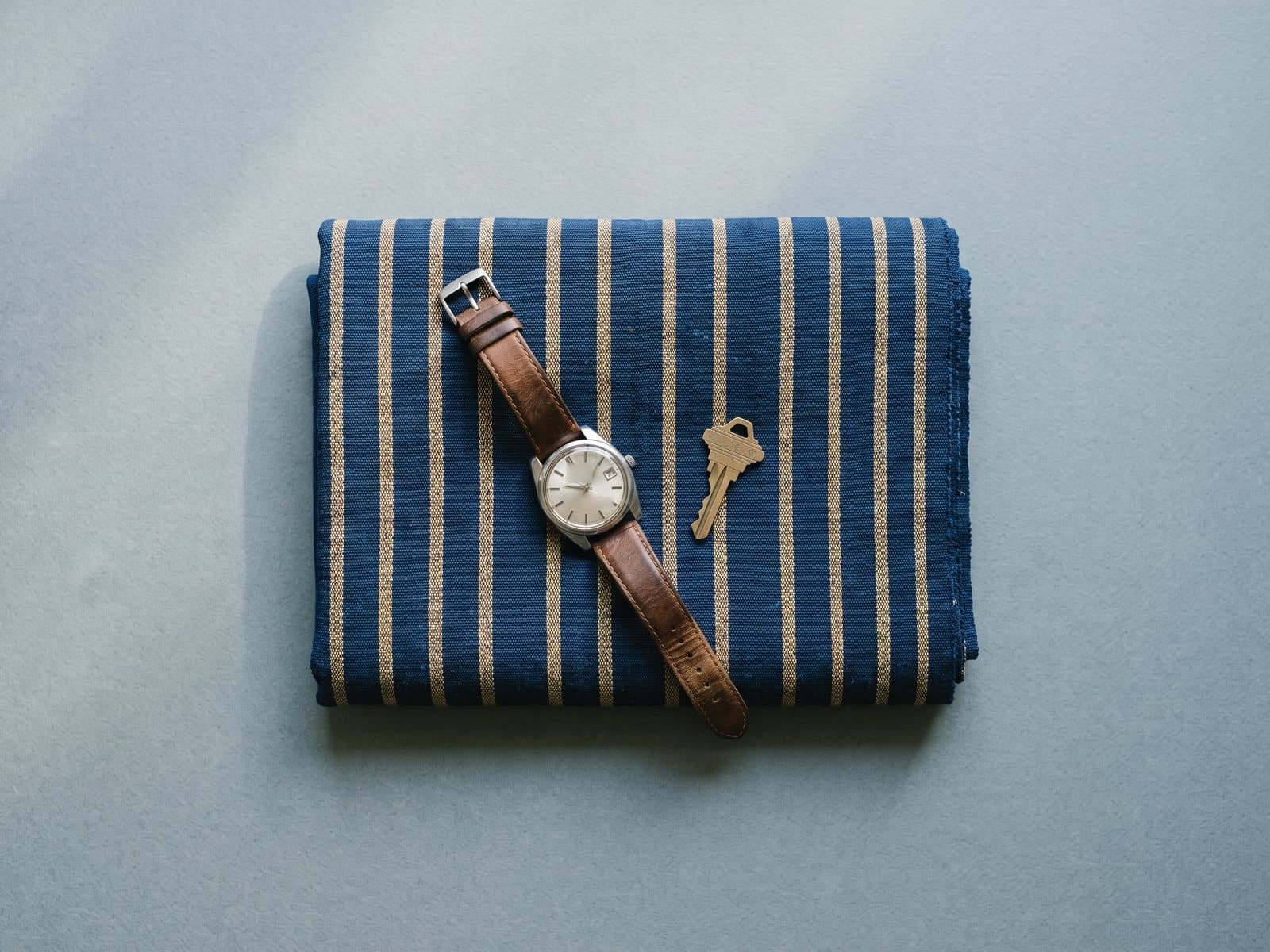 A folded length of indigo and gold aso-oke cloth with a wristwatch and a brass key on it.